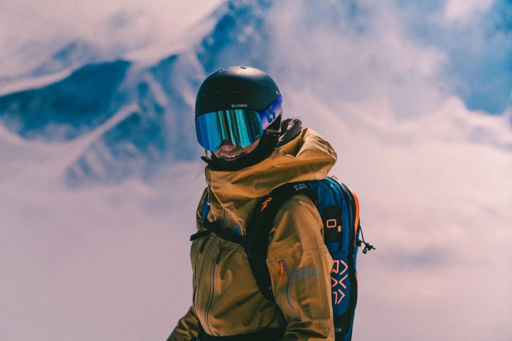 Skitourengeherin mit Helm, blau verspiegelter Skibrille, gelber Orage-Jacke und Rucksack vor Wolken und Bergen