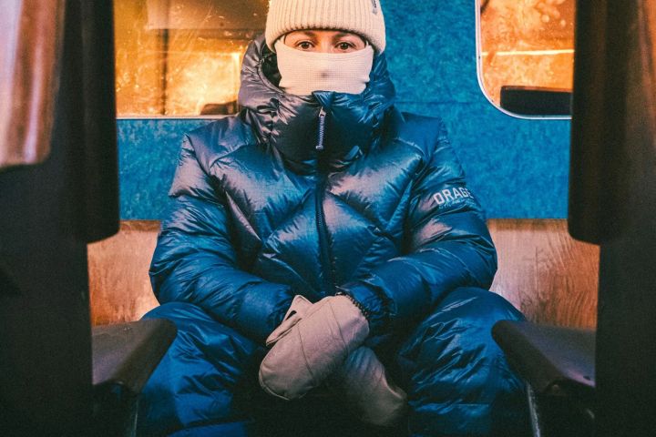 Person in glänzend blauer Daunenjacke mit hochgeschlossenem Kragen, Mütze und Fäustlingen sitzt in einem Zugabteil