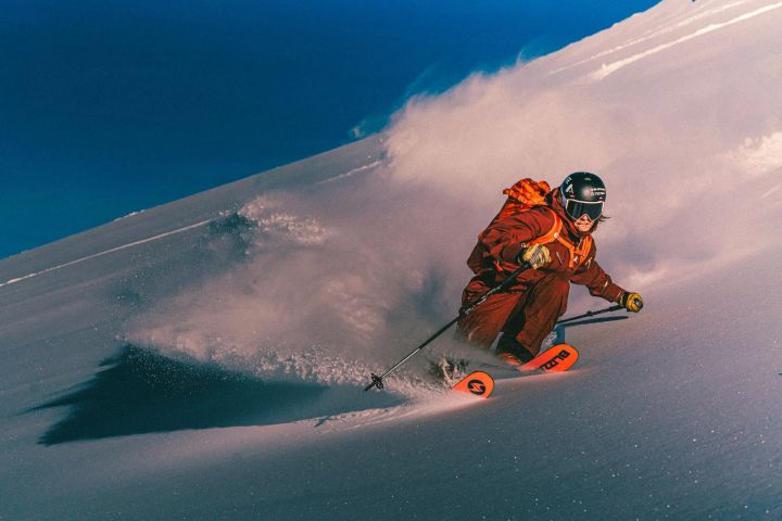 Skifahrer in orangefarbener Jacke und Hose zieht im Tiefschnee eine Schneefahne hinter sich her