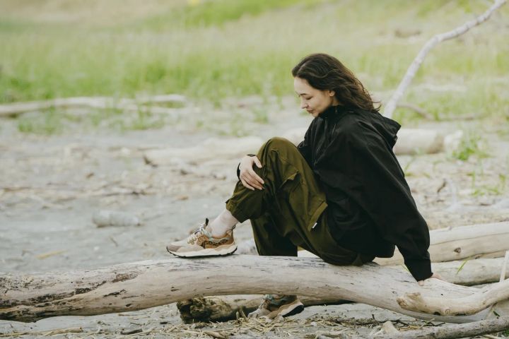 Frau in dunklem Kapuzenpullover und olivgrüner Hose sitzt auf einem Treibholzstamm am Strand, an den Füßen beige Sneaker