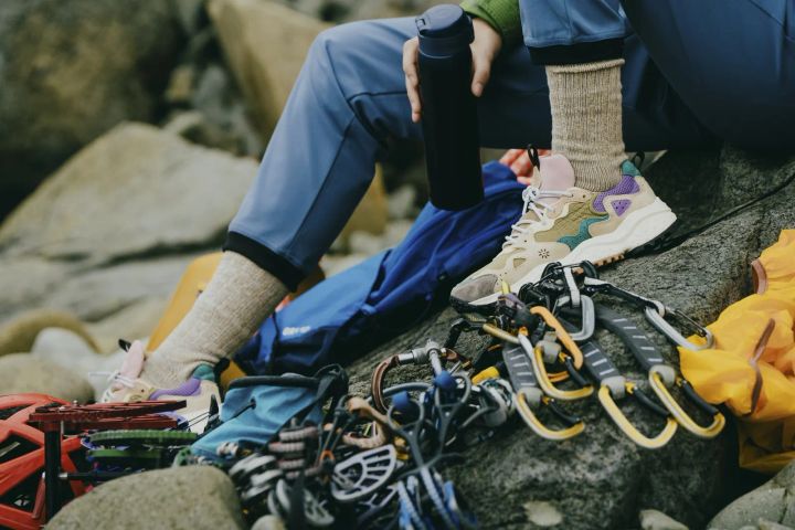 Füße in hellen Wollsocken und hellen Sneakern mit lila Details auf einem Felsen, daneben ein Kletterseil, Karabiner und eine Trinkflasche