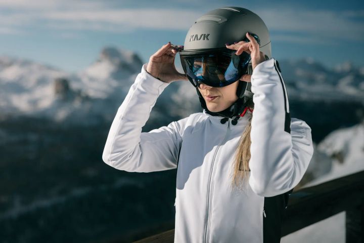 Frau in weißer Jacke rückt auf einer Terrasse vor verschneiten Gipfeln das Visier ihres grauen KASK-Helms zurecht