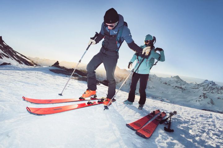 Zwei Skitourengeher mit Stöcken auf einem verschneiten Bergrücken, daneben ein abgelegtes Paar Ski