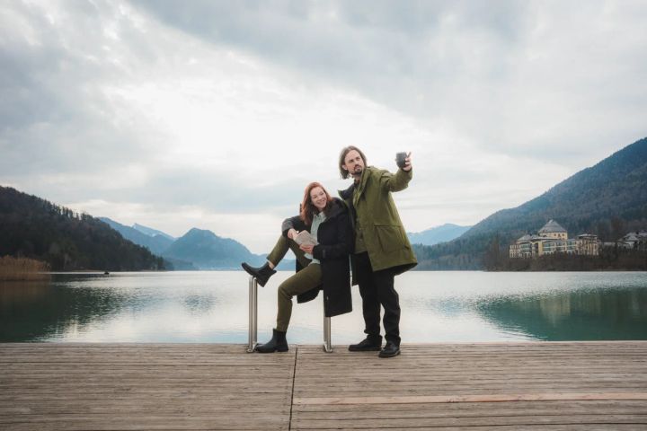 Frau und Mann in langen Mänteln stehen auf einem Holzsteg an einem Bergsee
