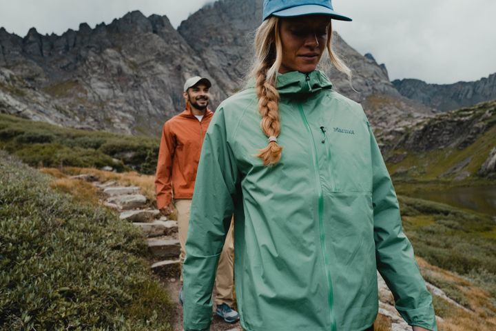 Wanderin in mintgrüner Jacke und Schirmmütze auf einem Bergpfad, hinter ihr ein Mann in orangefarbener Jacke