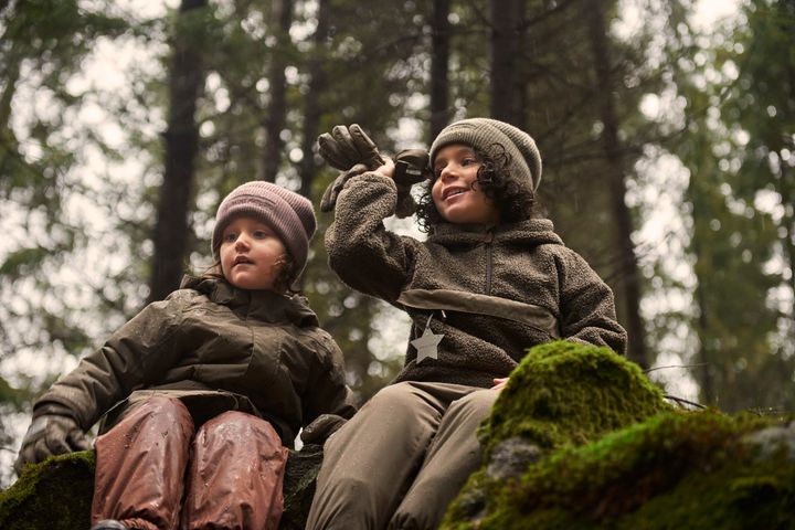 Zwei Kinder in braunen Fleecejacken und Mützen sitzen auf einem moosigen Felsen im Wald