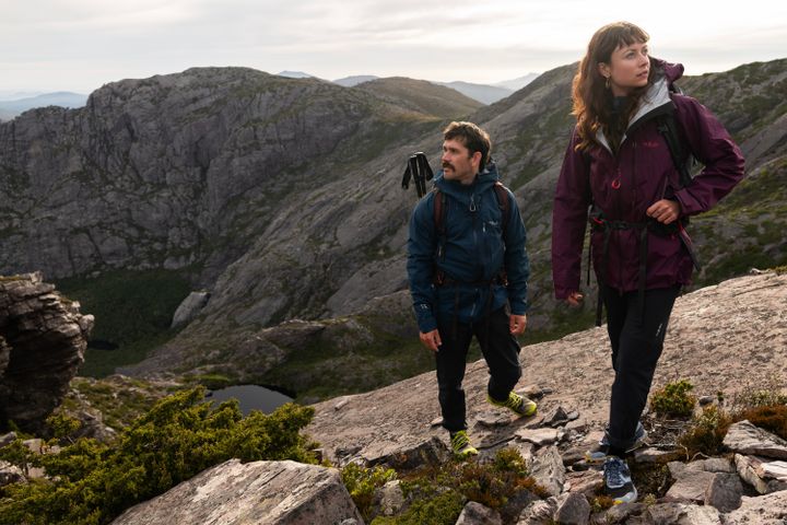Zwei Wandernde mit Rucksäcken auf einem felsigen Bergrücken, er in dunkelblauer, sie in weinroter Regenjacke, unten ein kleiner Bergsee