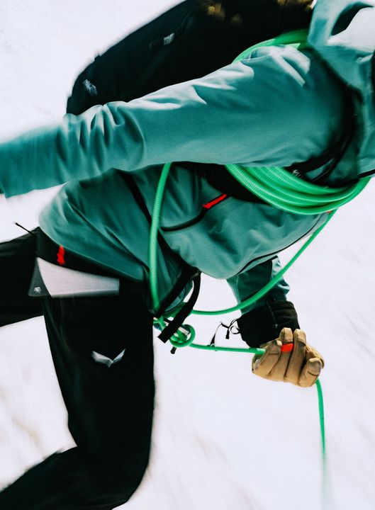 Bergsteiger in türkiser Jacke mit Rucksack, ein grünes Seil über der Schulter, vor verschneitem Hang