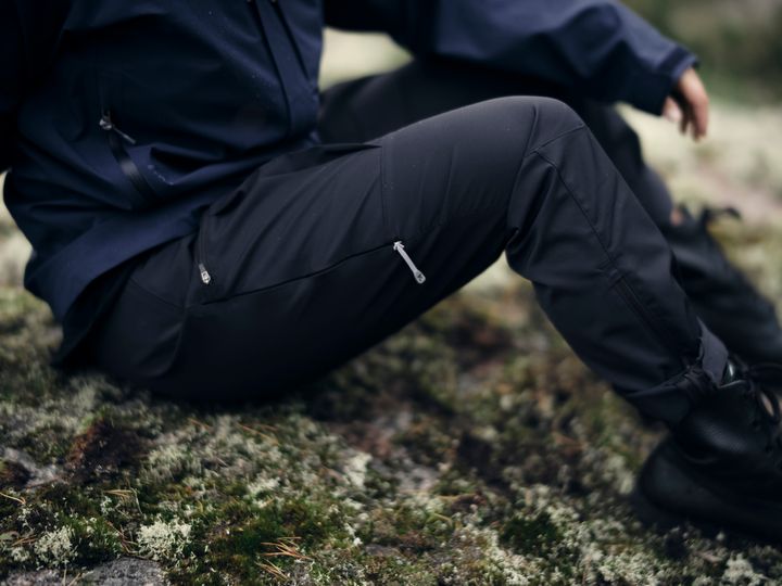 Person in dunkelblauer Outdoorhose und Jacke sitzt auf einem bemoosten Felsen