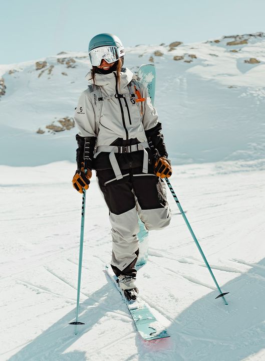Skifahrerin in hellgrau-schwarzer Jacke und Hose mit Helm und Skibrille geht mit Skiern und Stöcken über einen verschneiten Hang