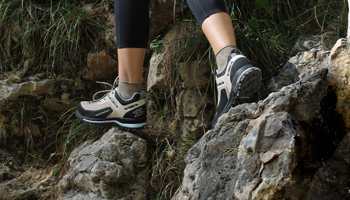 Wanderin in leichten, hellen Halbschuhen steigt auf einem felsigen, bewachsenen Pfad bergauf