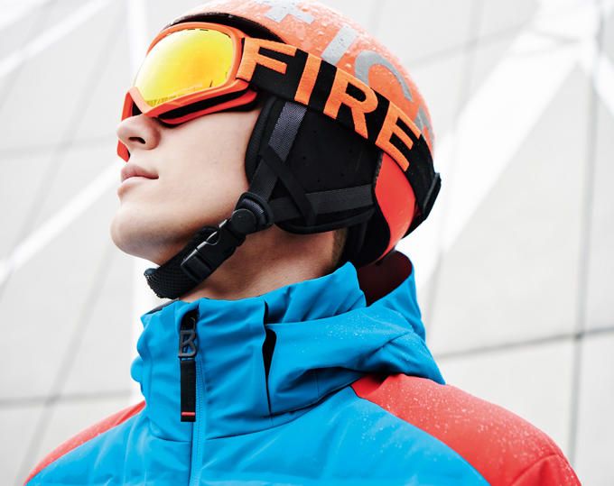 Mann mit orangem Skihelm und verspiegelter Skibrille in einer blau-roten Skijacke vor einer hellen Fassade