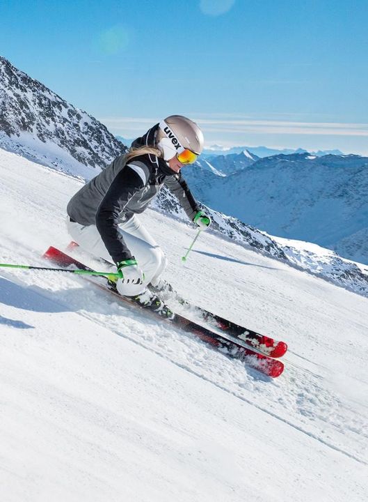 Skifahrerin mit hellem Uvex-Helm, orange verspiegelter Skibrille und grau-schwarzer Jacke fährt einen sonnigen Hang hinunter, dahinter verschneite Gipfel