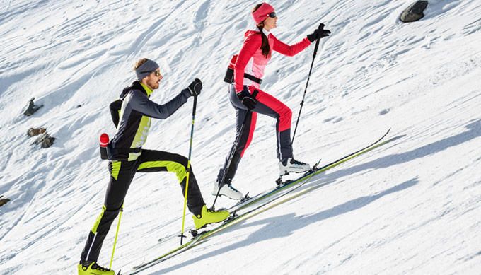 Langläuferin in Rot und Langläufer in Schwarz-Gelb steigen mit Stöcken auf schmalen Ski einen sonnigen Schneehang hinauf