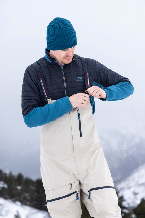 Mann in heller Latzhose mit Hosenträgern und blauer Jacke schließt im Schnee den Reißverschluss