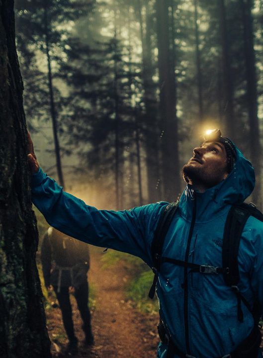 Wanderer in petrolblauer Regenjacke mit Kapuze und Stirnlampe stützt sich im nebligen Wald an einen Baumstamm, hinter ihm geht eine zweite Person auf dem Pfad