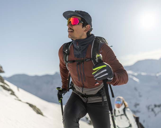 Skitourengeher mit Kappe, bunter Sportbrille und Rucksack steigt durch Schnee bergauf, die Hand im schwarz-gelben Handschuh am Stockgriff