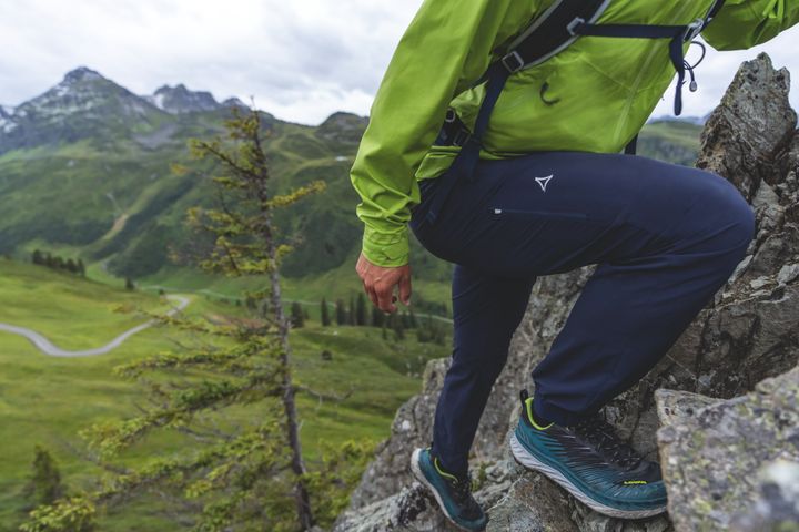 Wanderer in grüner Jacke und dunkelblauer Hose steigt mit hohem Schritt auf einen Felsen, dahinter Almwiesen und Berge