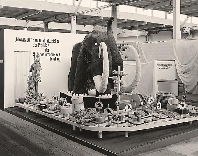 Historisches Schwarz-Weiß-Foto eines Messestands mit einer großen Mammut-Figur, davor ausgelegte Seile und Seilrollen, an der Wand der Schriftzug „Mammut“ als Qualitätszeichen