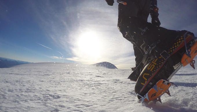 Bergschuh von La Sportiva mit angelegtem Steigeisen tritt in den Schnee, im Gegenlicht die Sonne