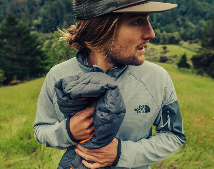 Mann mit Basecap in hellgrauer Fleecejacke von The North Face hält eine zusammengerollte graue Steppjacke vor der Brust, im Hintergrund Wiese und Wald