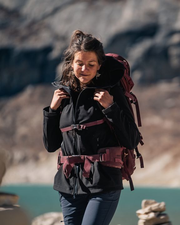 Wanderin mit dunkelrotem Trekkingrucksack greift an die Schulterriemen, Hüftgurt und Brustgurt sind geschlossen, im Hintergrund ein Bergsee