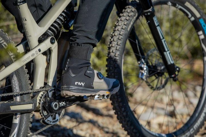 Fuß in grau-schwarzem Northwave-Radschuh mit hohem Schaft auf dem Pedal eines Mountainbikes, Waldboden im Hintergrund