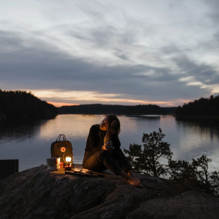 Frau sitzt in der Dämmerung auf einem Felsen über einem See, neben ihr leuchten eine Gaslaterne und eine kleine Lampe
