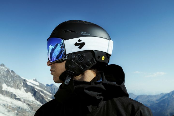 Mann im Profil mit schwarzem Sweet-Protection-Helm und blau verspiegelter Skibrille mit weißem Band vor verschneiten Gipfeln und blauem Himmel