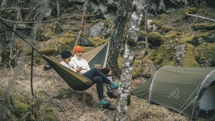 Zwei Personen in Pullovern sitzen in einer olivfarbenen Hängematte zwischen Bäumen im Wald, daneben steht ein Zelt