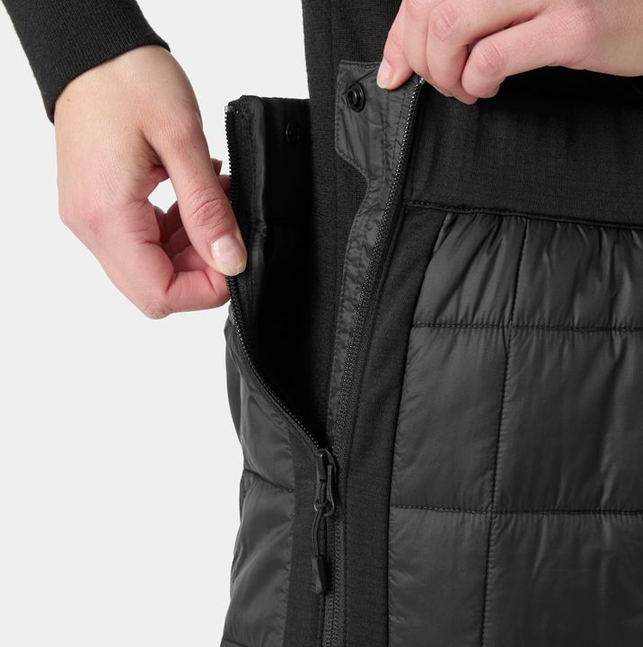 Hand öffnet den seitlichen Reißverschluss einer schwarzen, gesteppten Isolationshose von Helly Hansen