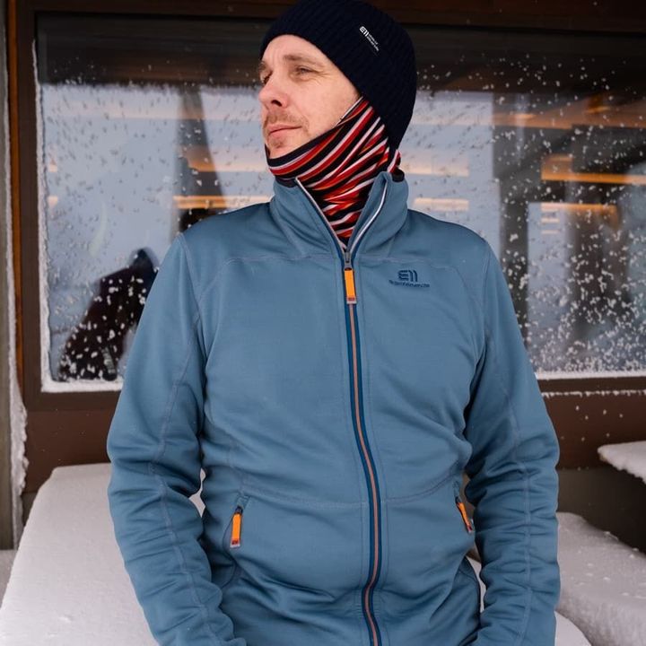 Mann in blauer Fleecejacke mit Mütze und gestreiftem Halstuch vor einer verschneiten Hütte