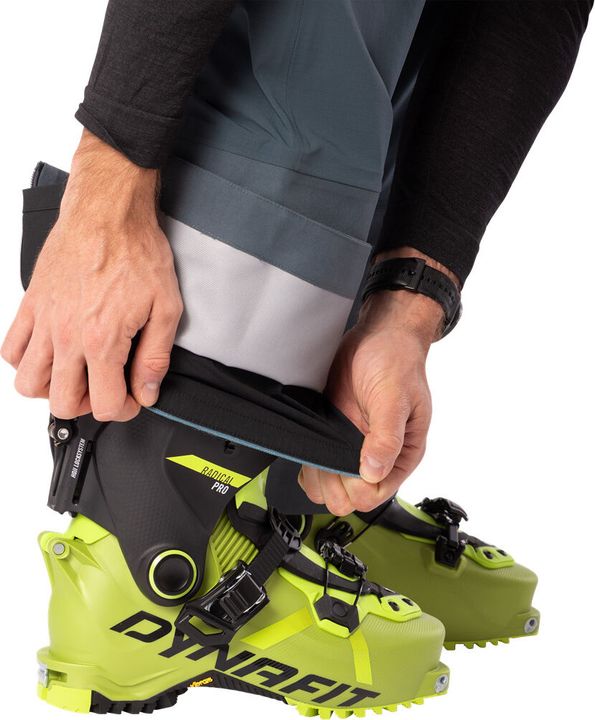 Hand öffnet den Reißverschluss am Beinabschluss einer grauen Dynafit-Skitourenhose über einem gelben Tourenskischuh