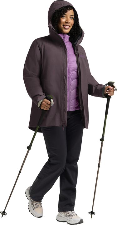 Frau mit Wanderstöcken in auberginefarbener Jacke mit Kapuze, darunter eine fliederfarbene Daunenjacke