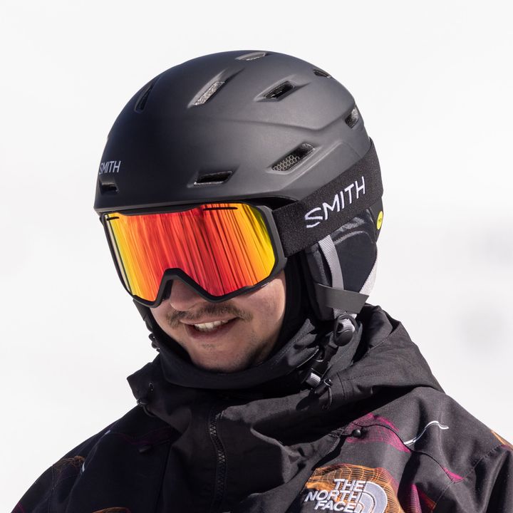 Lächelnder Mann mit schwarzem Smith-Helm und rot-orange verspiegelter Skibrille, der Rahmen liegt ohne Spalt am Helm an
