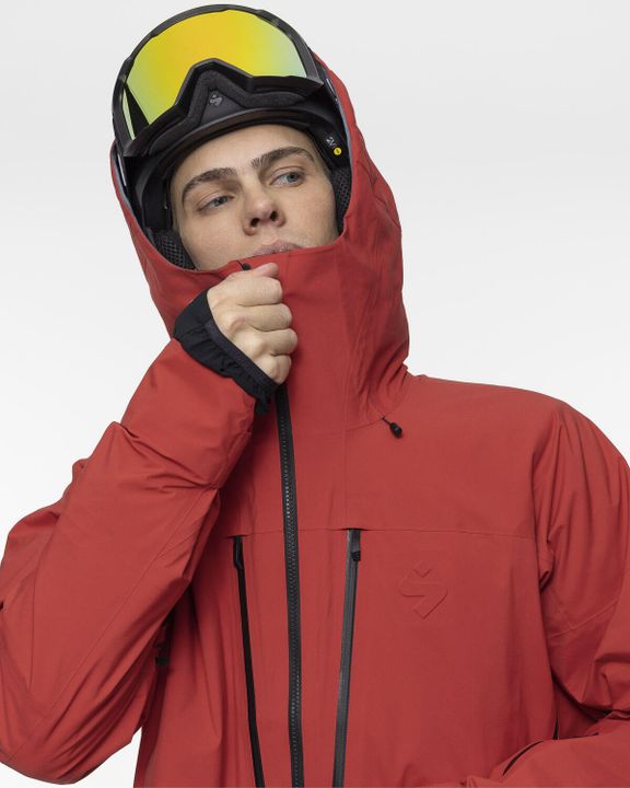Mann in roter Skijacke, die Kapuze über dem schwarzen Helm, die gelb verspiegelte Skibrille hochgeschoben, zieht den Kragen bis zur Nase hoch