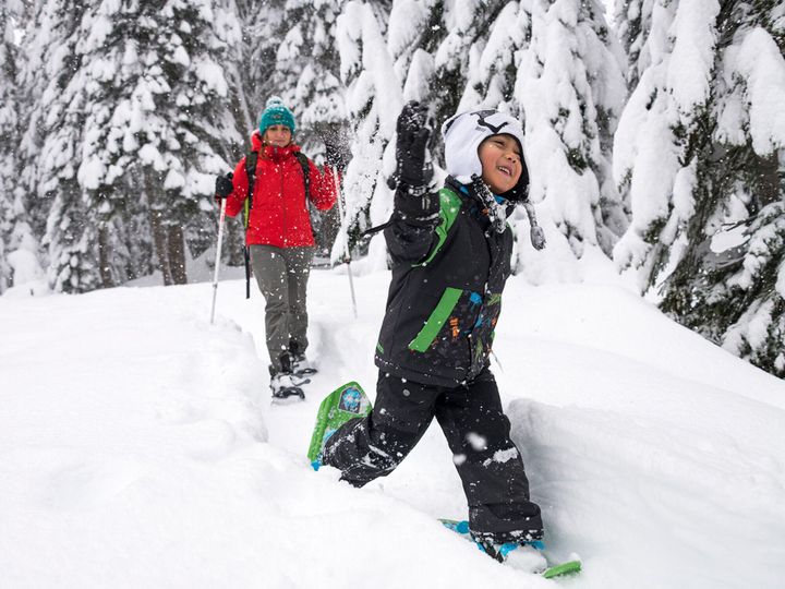Kind mit grünen Schneeschuhen läuft lachend durch tiefen Schnee im verschneiten Wald, dahinter eine Frau mit Stöcken