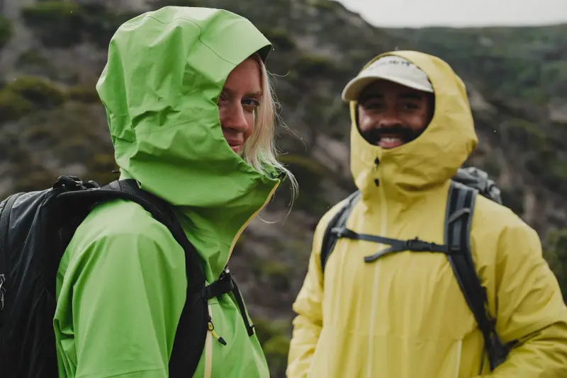 Zwei Personen mit Rucksäcken und geschlossenen Kapuzen an grüner und gelber Regenjacke