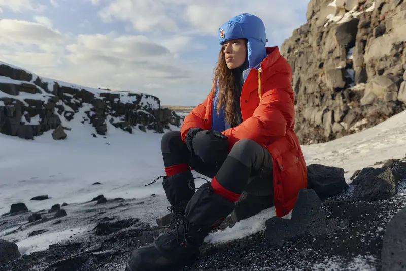 Frau in roter Daunenjacke mit blauer Mütze auf verschneitem Fels