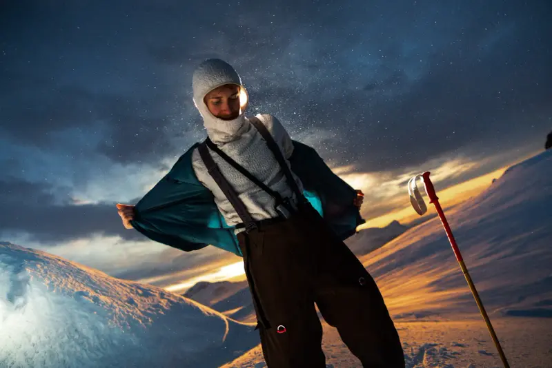 Person mit Sturmhaube und Skilatzhose öffnet die Jacke im Abendlicht auf einem verschneiten Hang